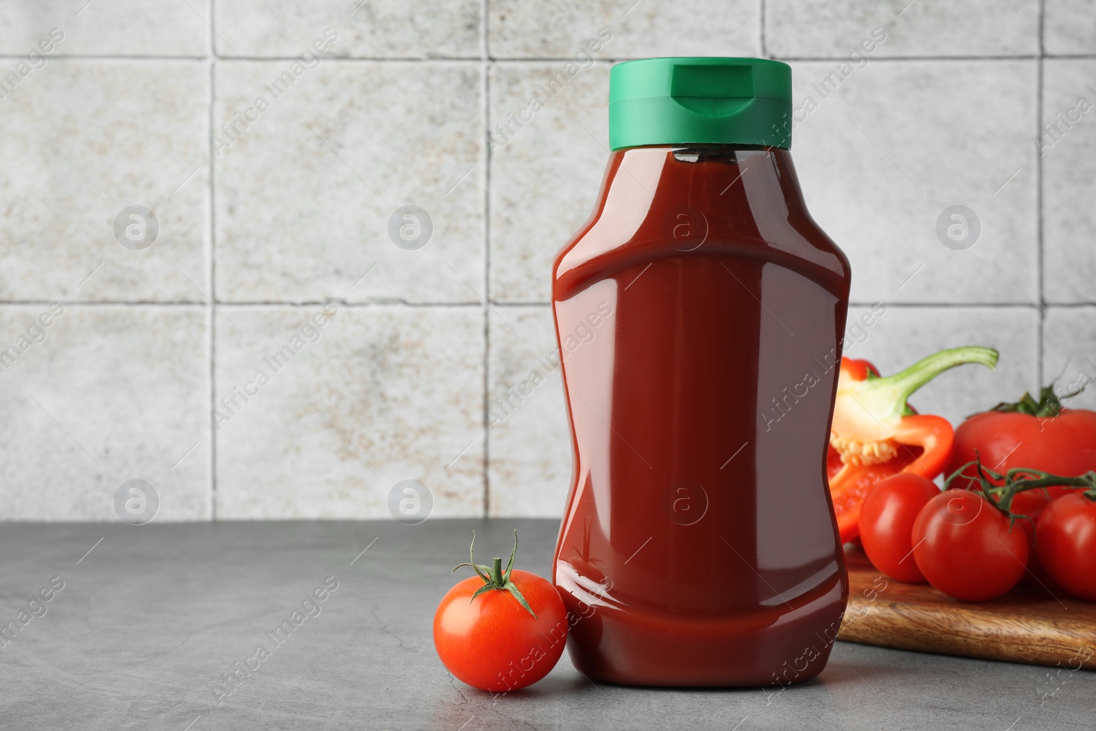 Photo of Bottle of tasty ketchup, tomatoes and pepper on grey table. Space for text