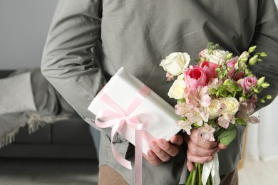 Man hiding bouquet of flowers and present indoors, closeup