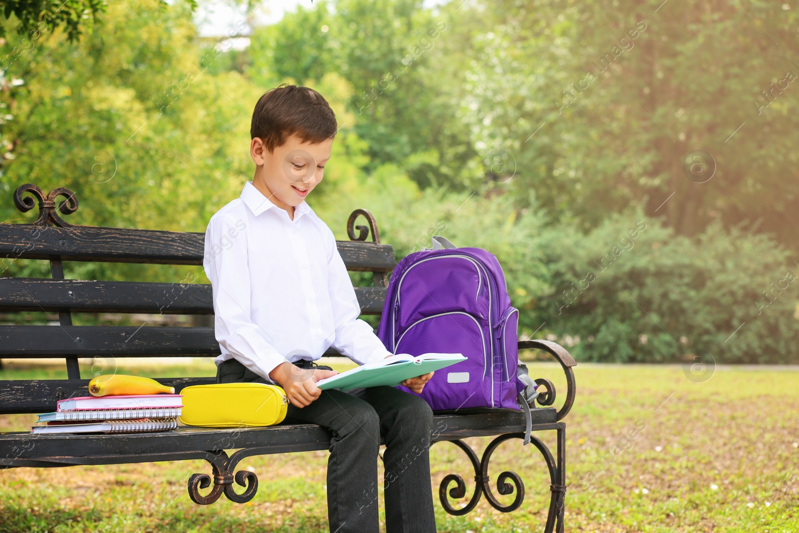 Photo of Cute little school child with stationery reading book on bench in park