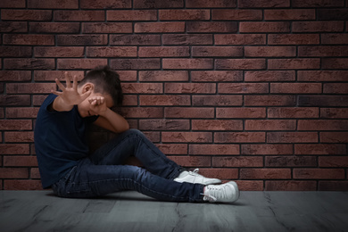 Photo of Scared little boy closing eyes with hand on floor near brick wall. Child in danger