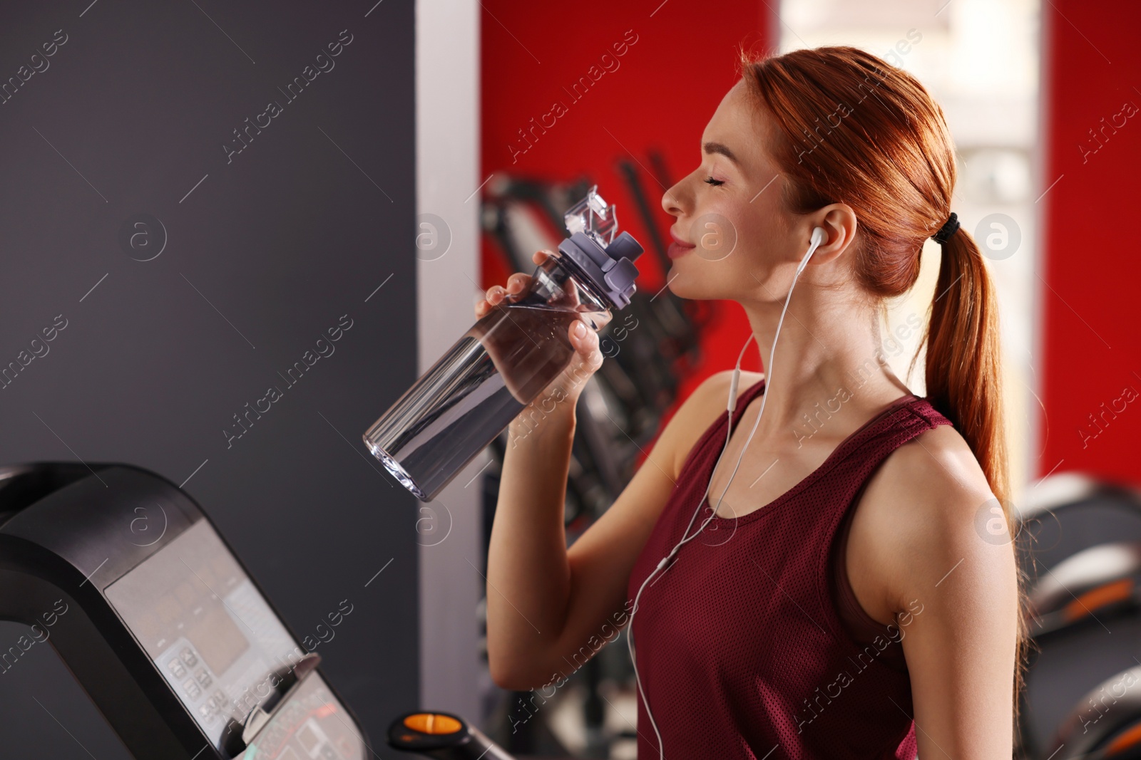 Photo of Athletic young woman with earphones and bottle of water training on treadmill in gym