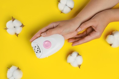 Woman holding modern epilator near fluffy cotton flowers on yellow background, top view
