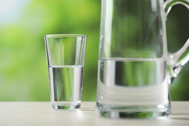 Photo of Jug and glass with clear water on white table against blurred green background, closeup