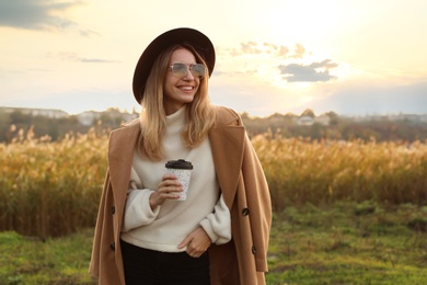 Beautiful young woman with cup of coffee wearing stylish autumn clothes outdoors