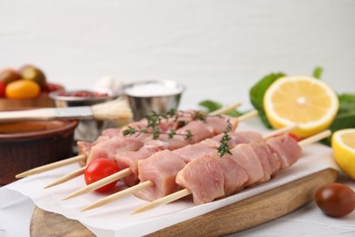 Skewers with cut raw meat, thyme and tomatoes on light table, closeup