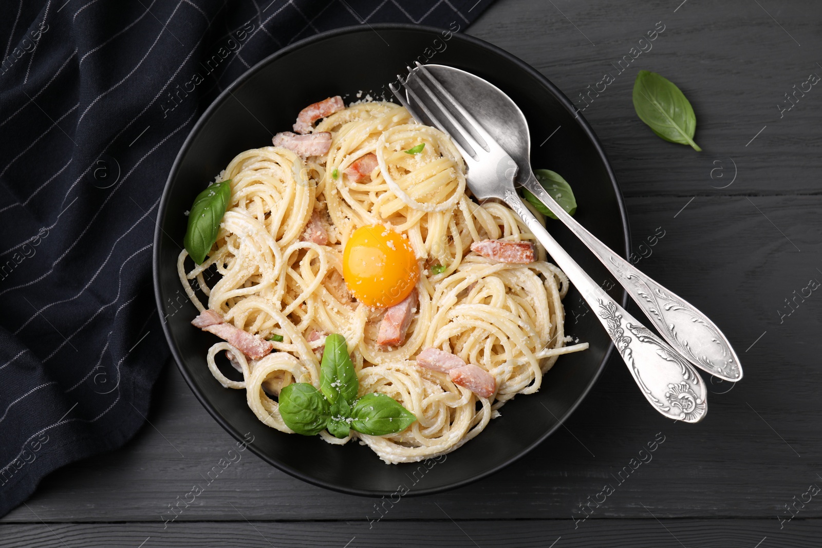 Photo of Bowl of delicious pasta Carbonara with egg yolk and cutlery on black wooden table, flat lay