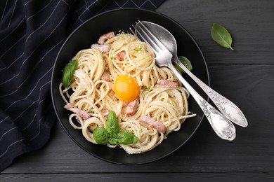 Photo of Bowl of delicious pasta Carbonara with egg yolk and cutlery on black wooden table, flat lay