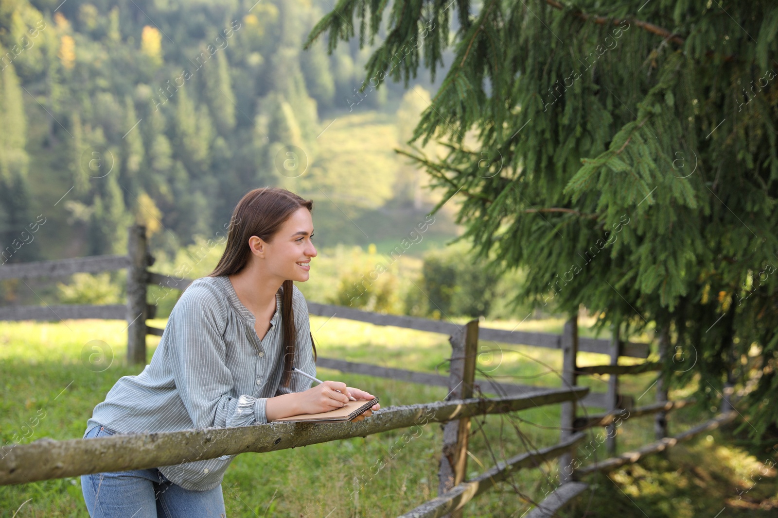 Photo of Beautiful young woman drawing with pencil in notepad near wooden fence