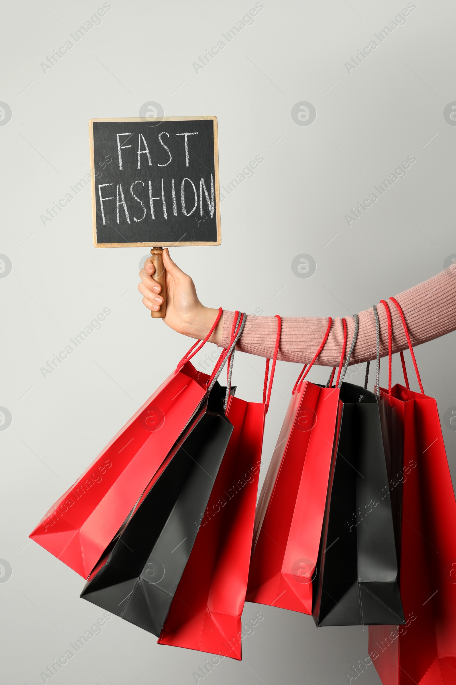 Photo of Woman holding small chalkboard with phrase Fast Fashion and different shopping bags on light background, closeup