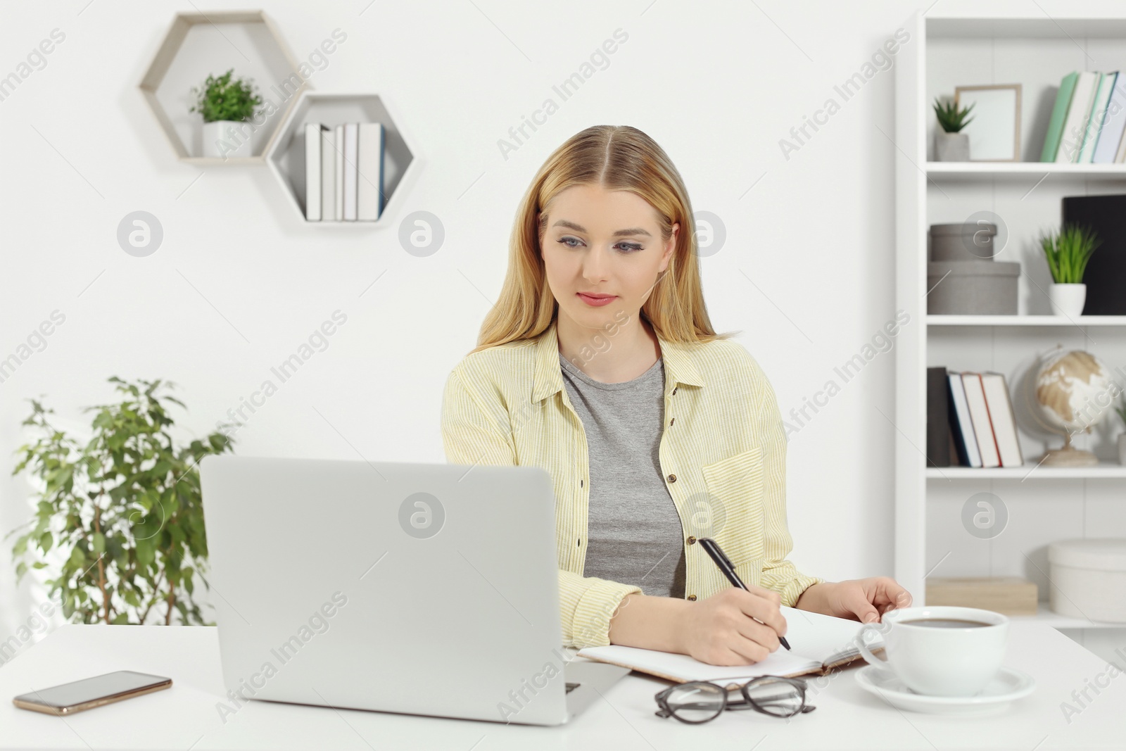 Photo of Home workplace. Woman writing in notebook near laptop at white desk in room