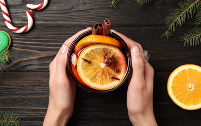 Photo of Woman holding glass cup of tasty mulled wine at black wooden table, top view