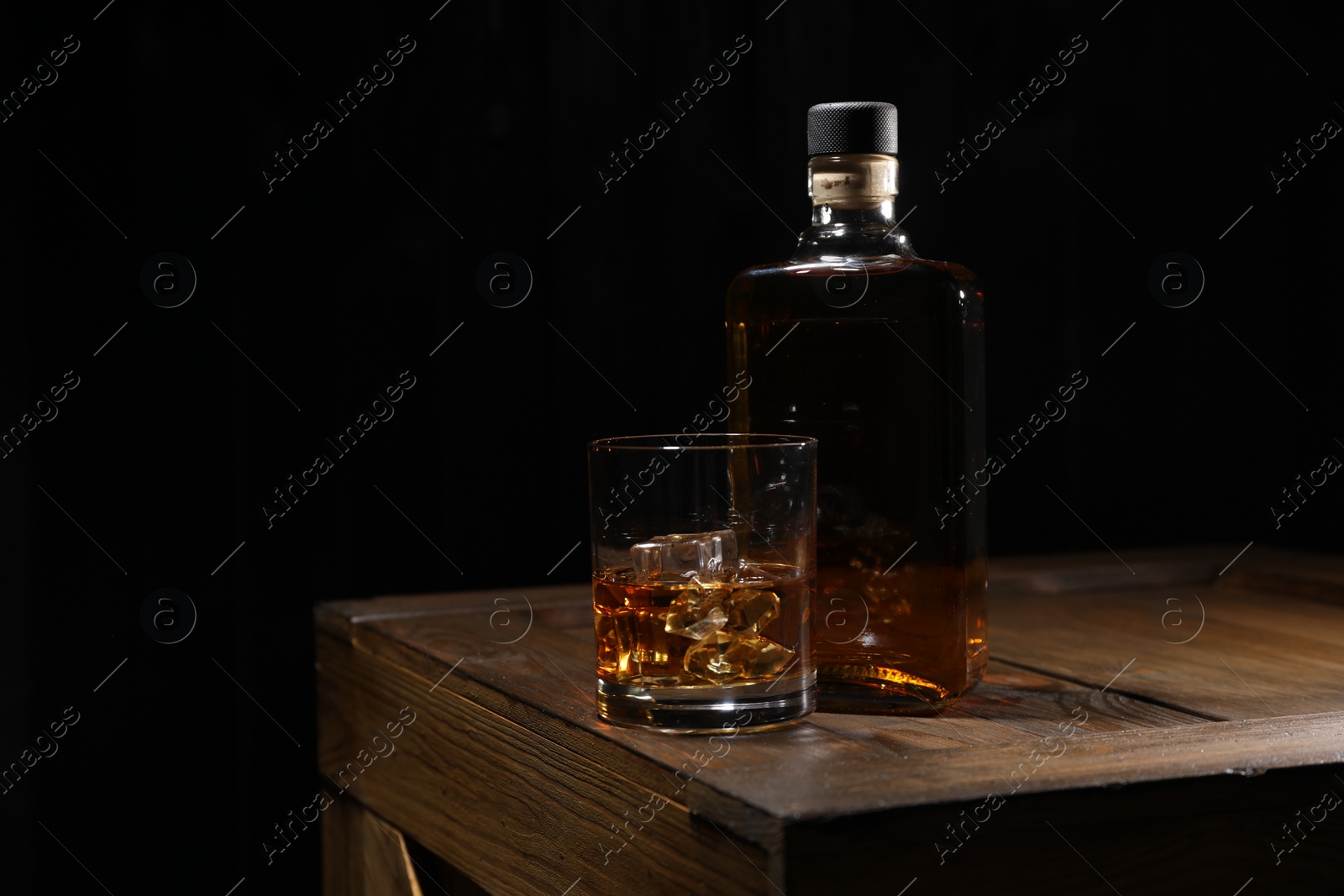 Photo of Whiskey with ice cubes in glass and bottle on wooden crate against black background