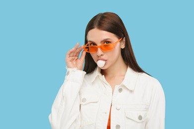 Beautiful woman in sunglasses blowing bubble gum on light blue background