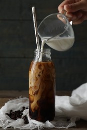 Woman pouring milk into bottle with iced coffee at wooden table, closeup