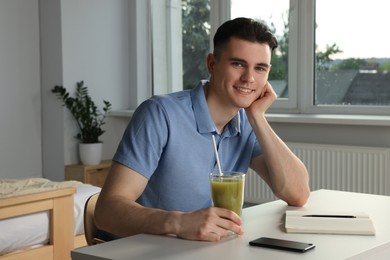Photo of Handsome young man with glass of juice sitting at table in bedroom