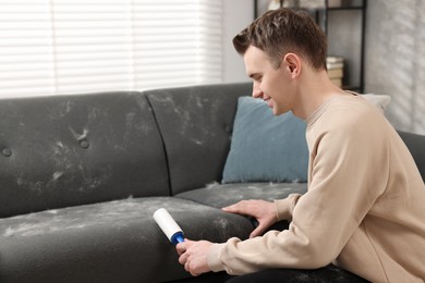 Photo of Pet shedding. Smiling man with lint roller removing dog's hair from sofa at home