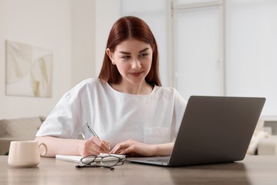 Photo of Beautiful woman using laptop and writing notes at wooden table indoors