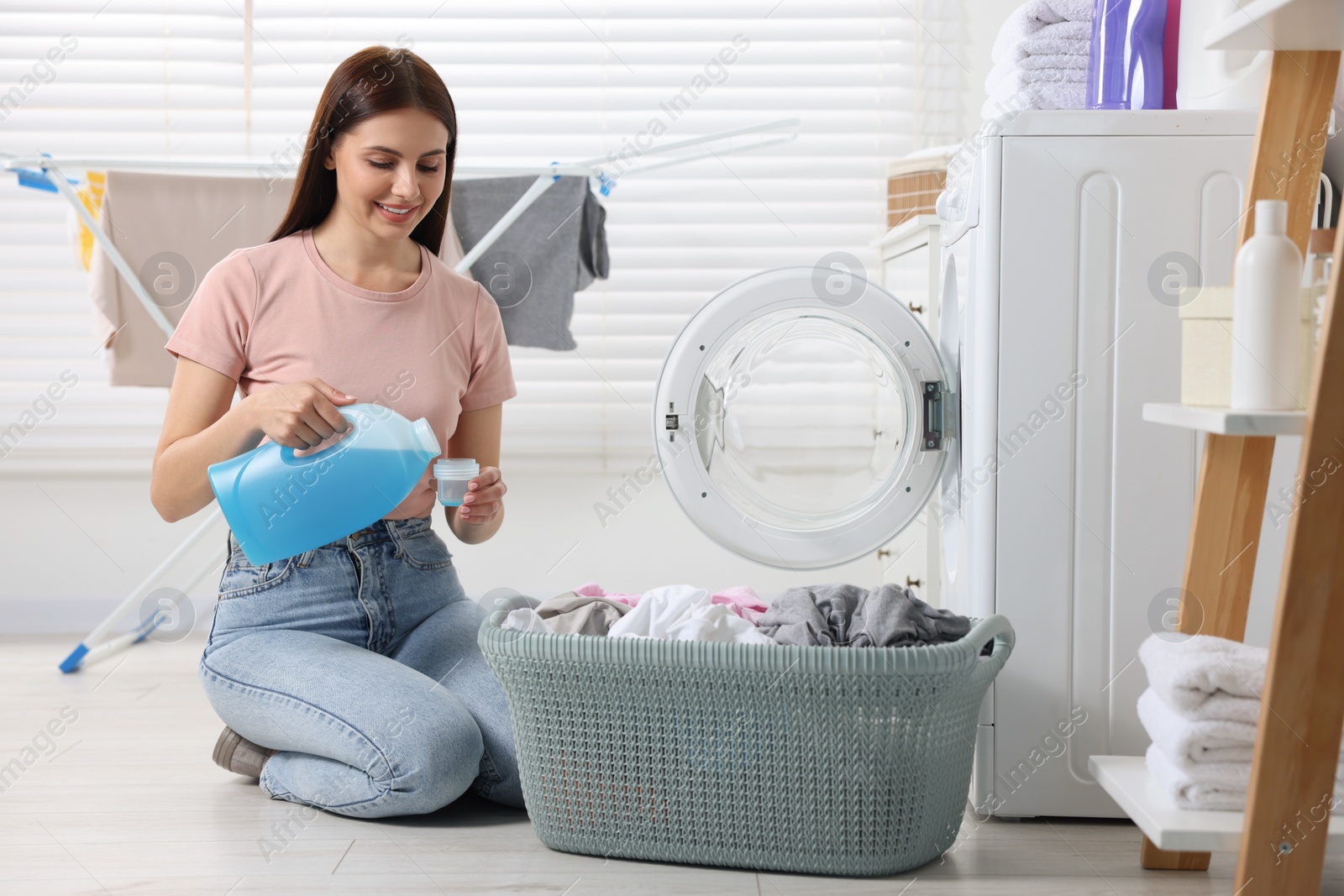 Photo of Woman pouring fabric softener from bottle into cap near washing machine in bathroom