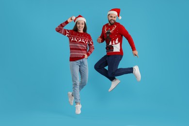 Photo of Happy young couple in Christmas sweaters and Santa hats jumping on light blue background
