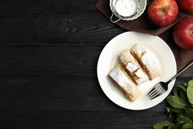 Photo of Delicious strudel, powdered sugar and fresh apples on black wooden table, flat lay. Space for text