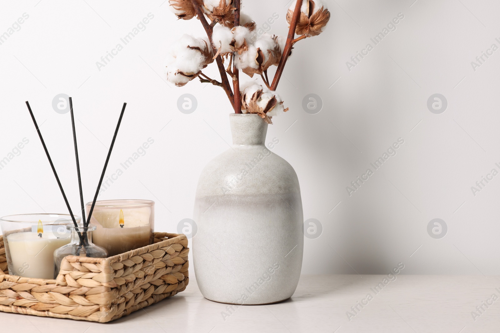 Photo of Burning candles in wicker basket and vase with cotton branches on table against white background. Space for text