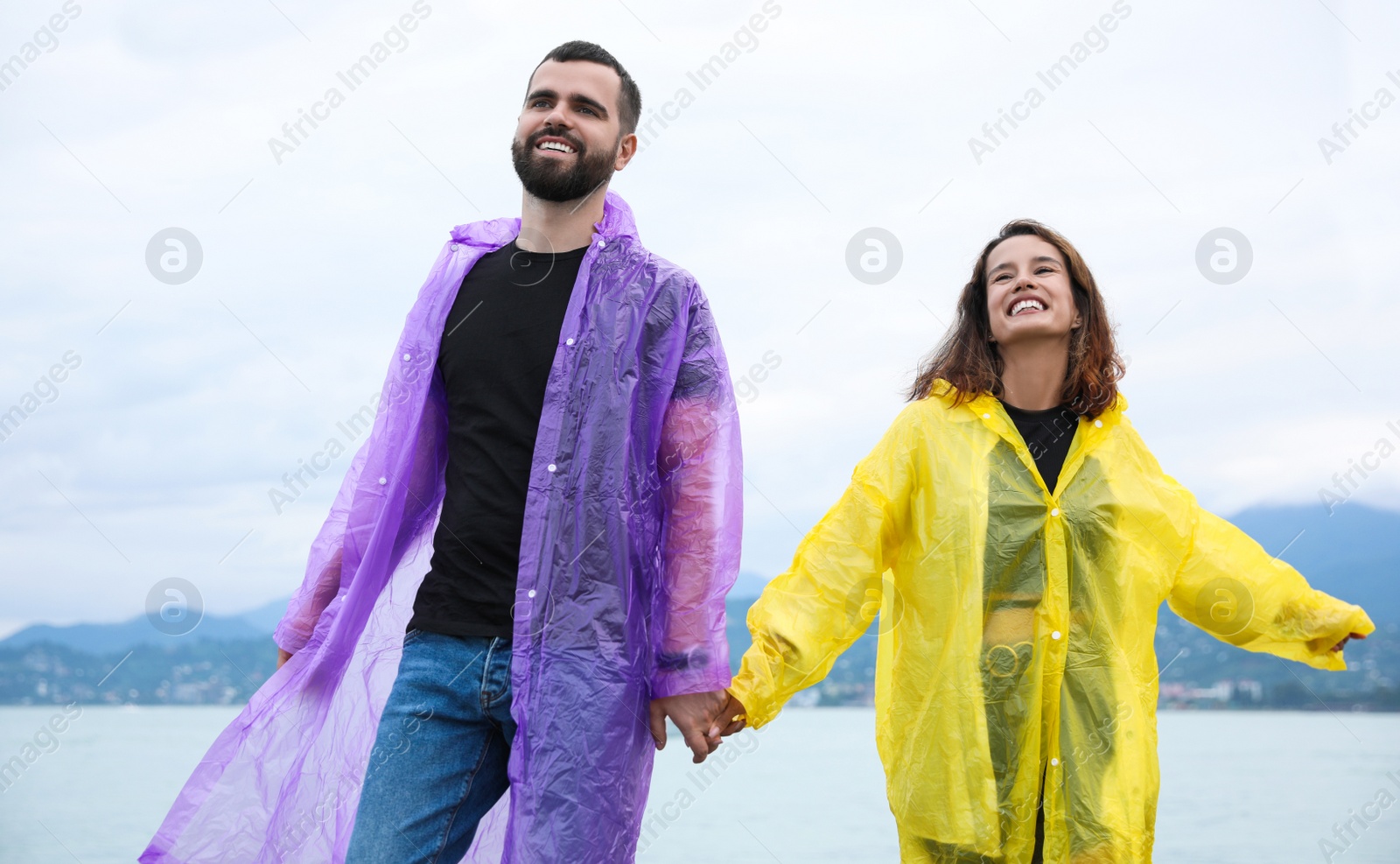 Photo of Young couple in raincoats enjoying time together under rain on beach