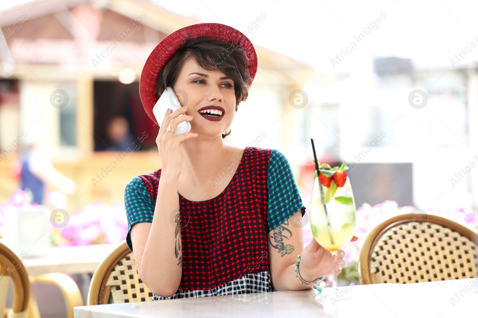 Photo of Young woman with glass of tasty lemonade talking on phone in open-air cafe