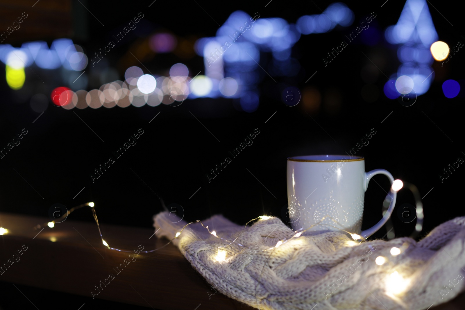 Photo of Cup of hot beverage, sweater and Christmas lights on wooden railing outdoors against blurred background, space for text. Winter night