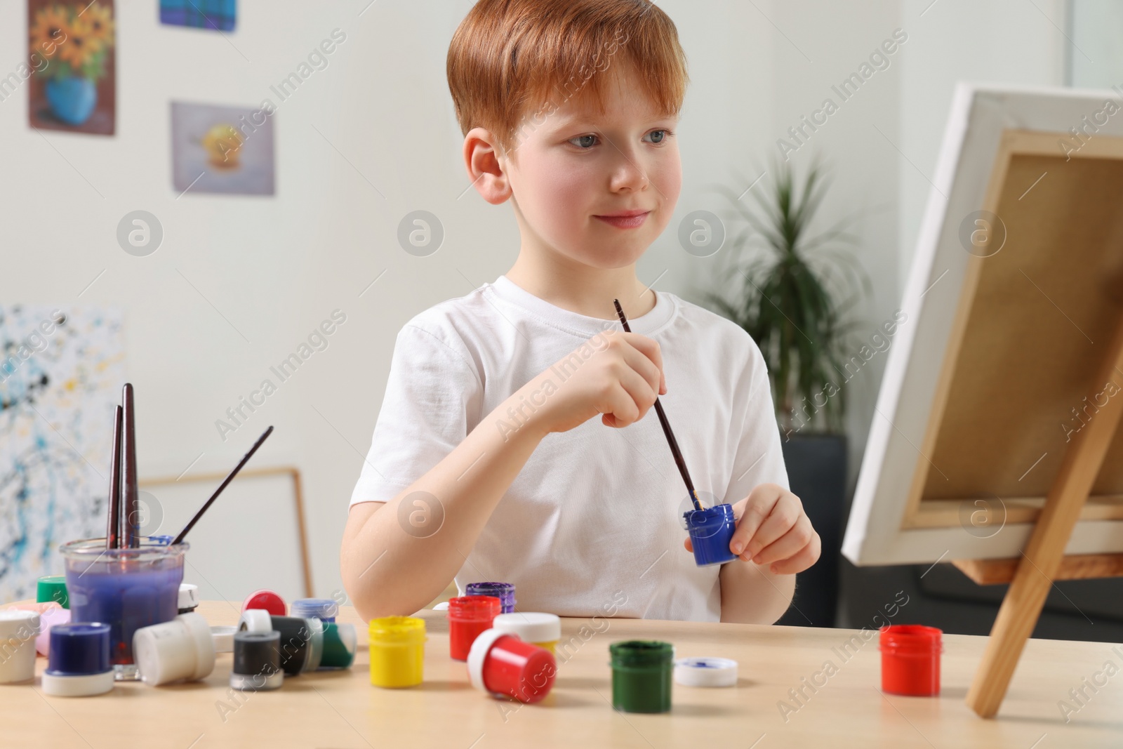 Photo of Little boy painting at table in studio. Using easel to hold canvas