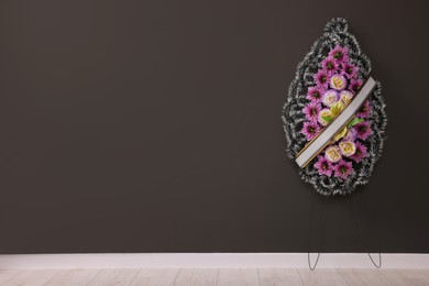 Funeral wreath of plastic flowers with ribbon hanging on dark grey wall indoors, space for text