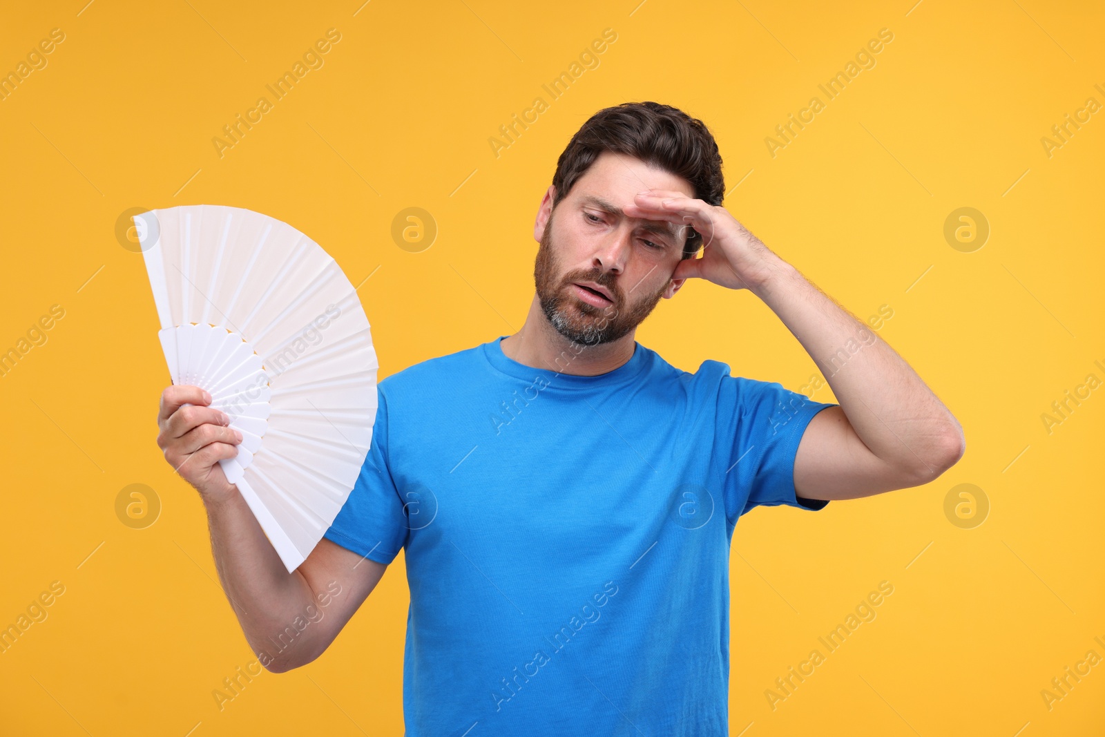 Photo of Unhappy man with hand fan suffering from heat on orange background