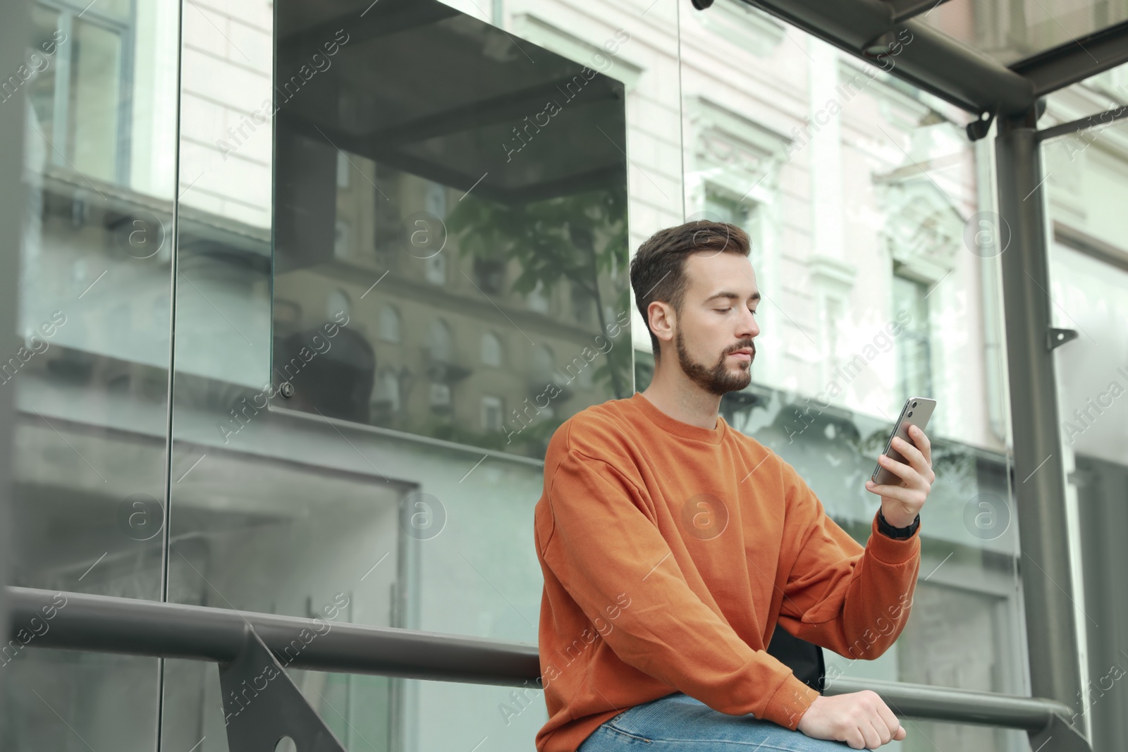 Photo of Young man with smartphone waiting for public transport at bus stop