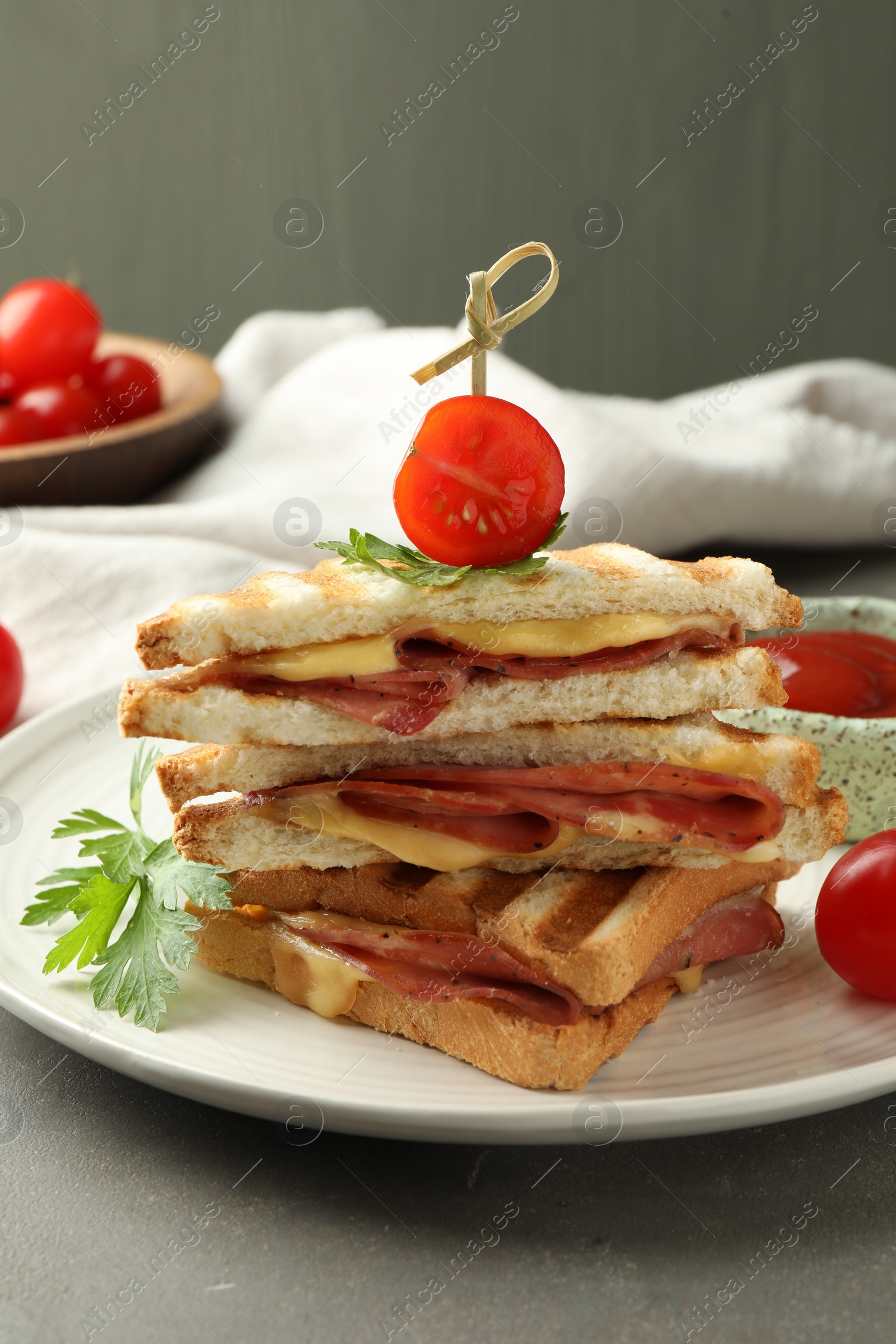 Photo of Stack of tasty sandwiches with ham and melted cheese served with tomato on grey textured table, closeup