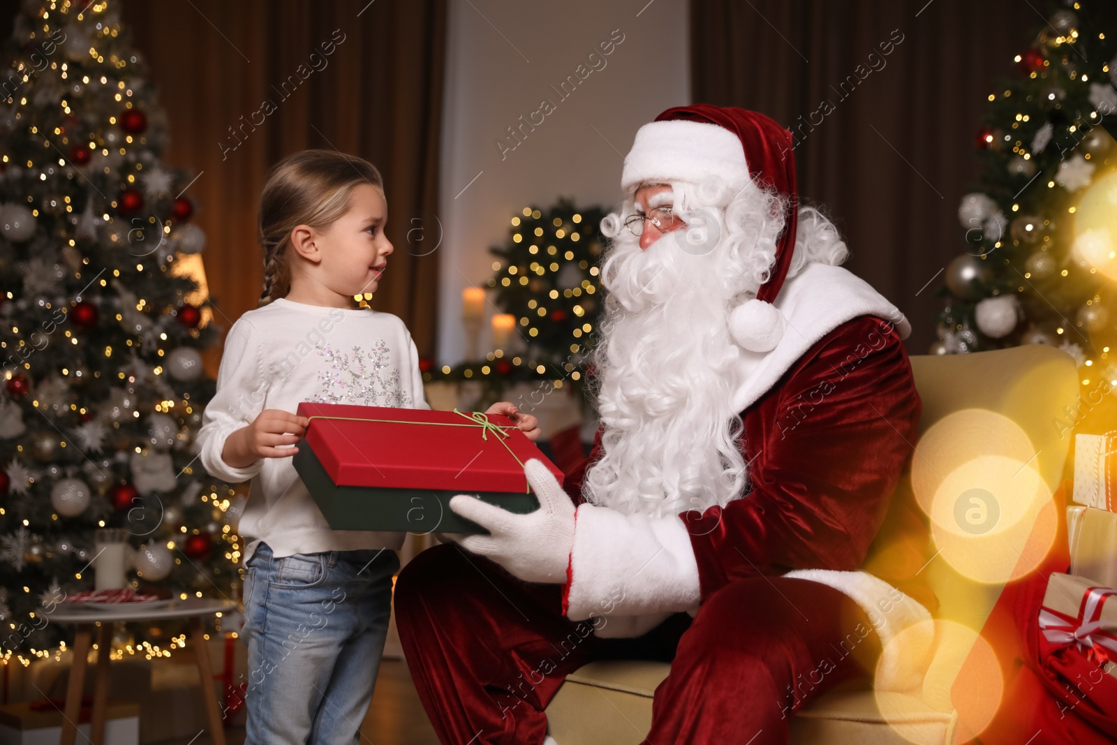 Photo of Santa Claus giving present to little girl in room decorated for Christmas