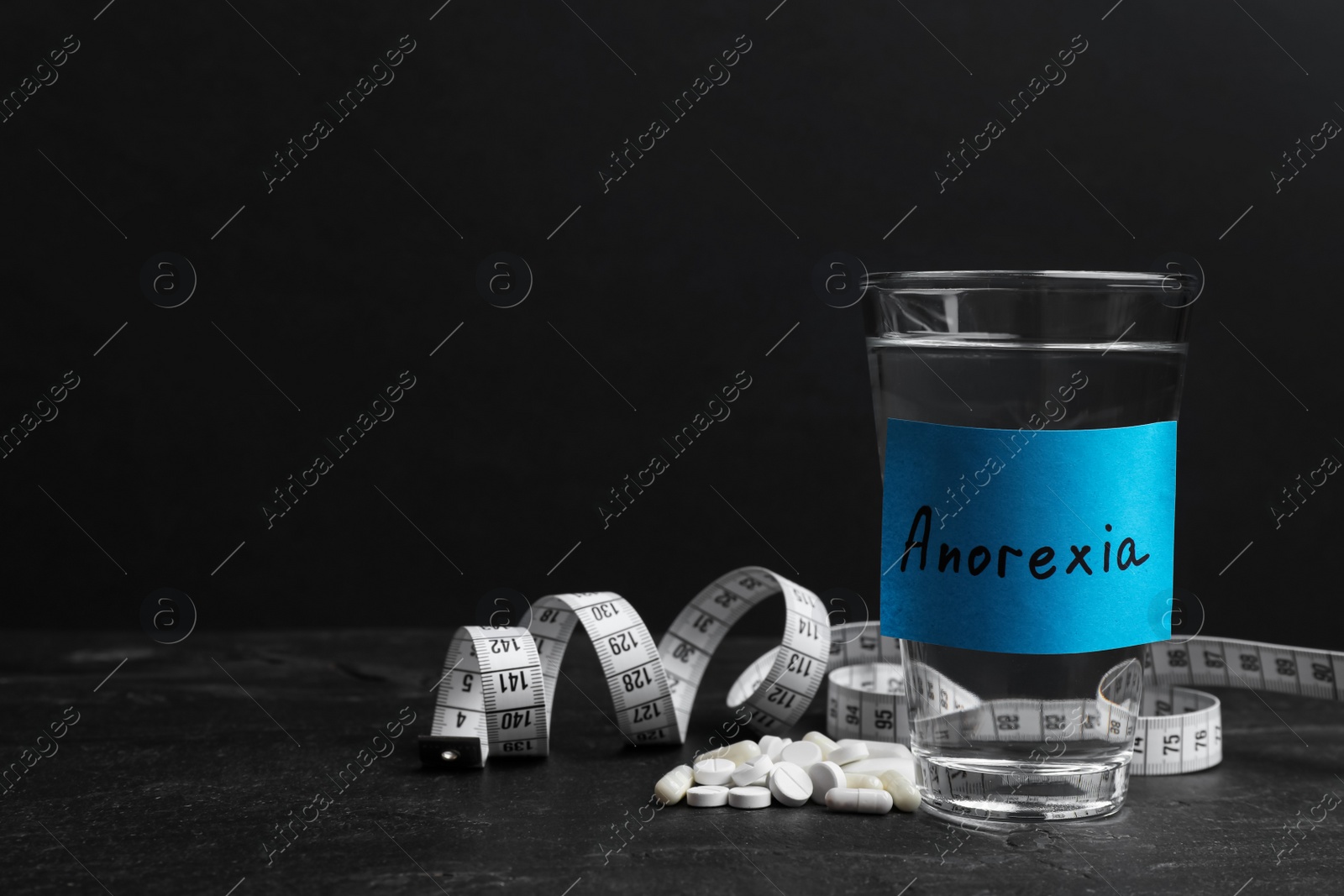 Photo of Sticky note with word Anorexia attached to glass of water near pills and measuring tape on black table, space for text