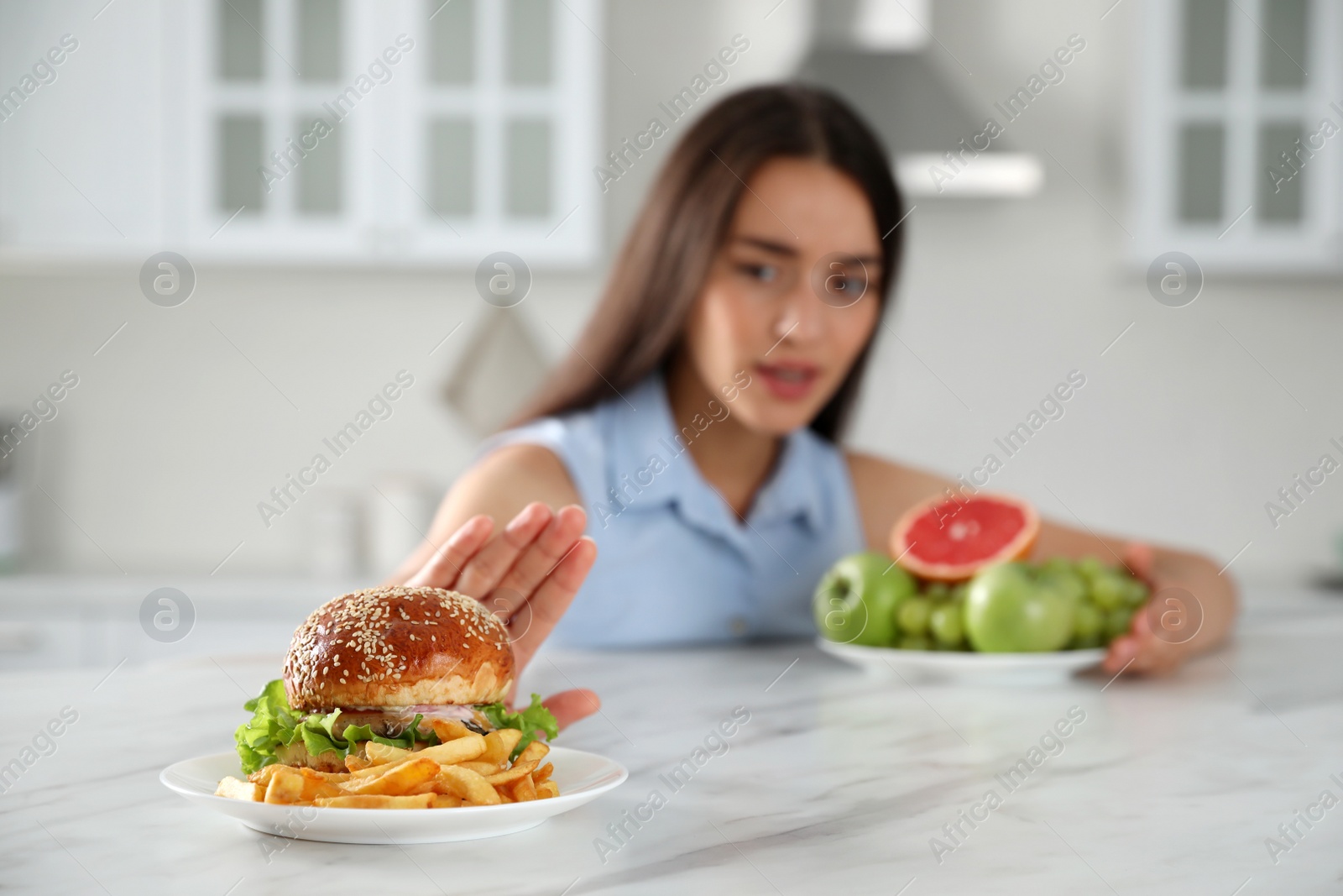 Photo of Woman choosing between fruits and burger with French fries in kitchen, focus on food