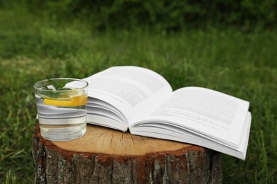 Photo of Open book near glass of water with mint and lemon on tree stump outdoors