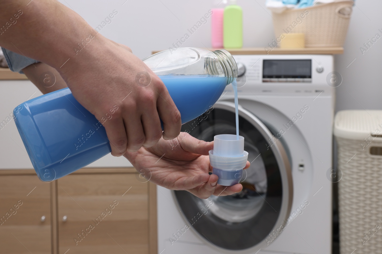 Photo of Man pouring fabric softener from bottle into cap near washing machine indoors, closeup