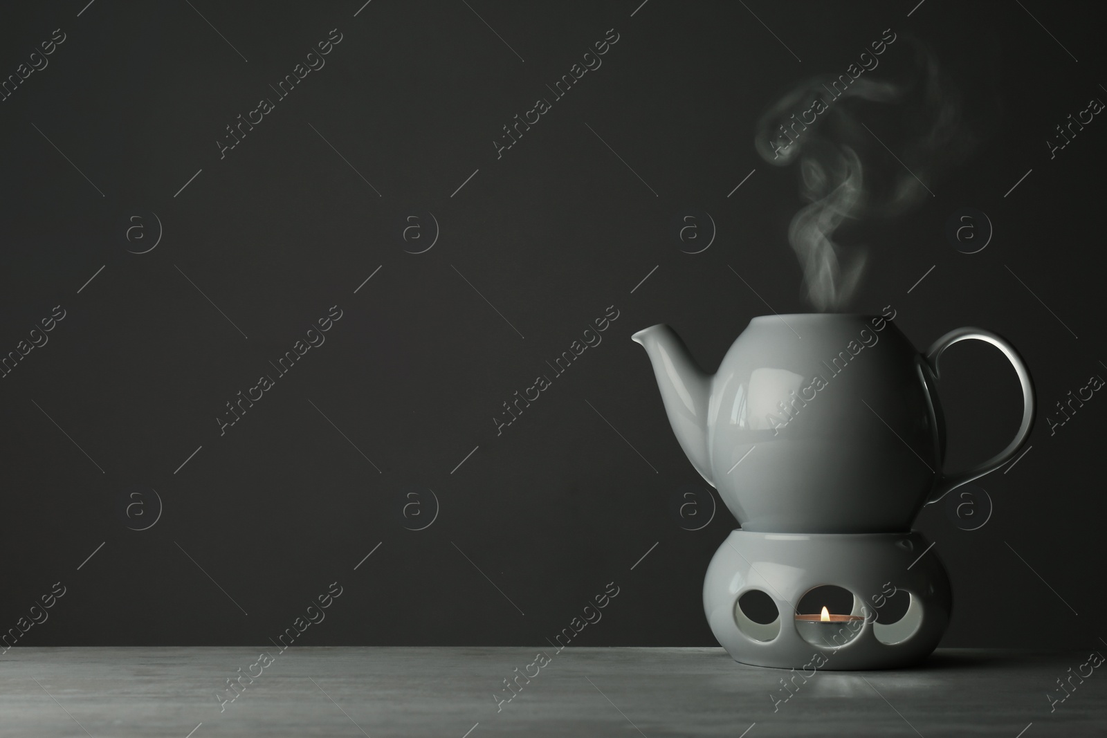 Photo of Ceramic teapot and warmer with burning candle on table against grey background. Space for text