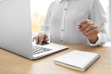 Woman with notebook and pen working on laptop at wooden table, closeup. Electronic document management