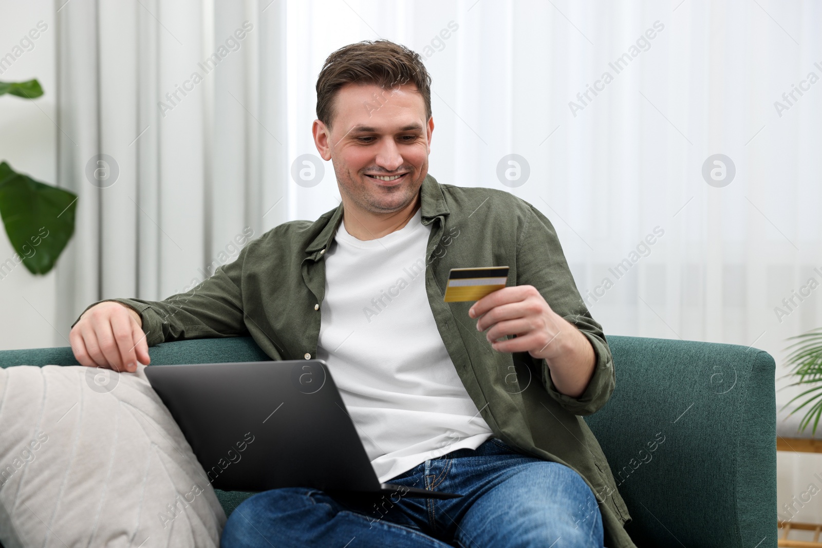 Photo of Happy man with credit card using laptop for shopping online on sofa at home