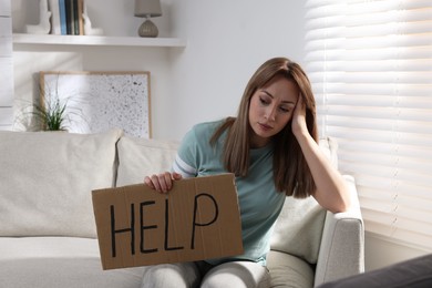 Unhappy young woman with HELP sign on sofa indoors