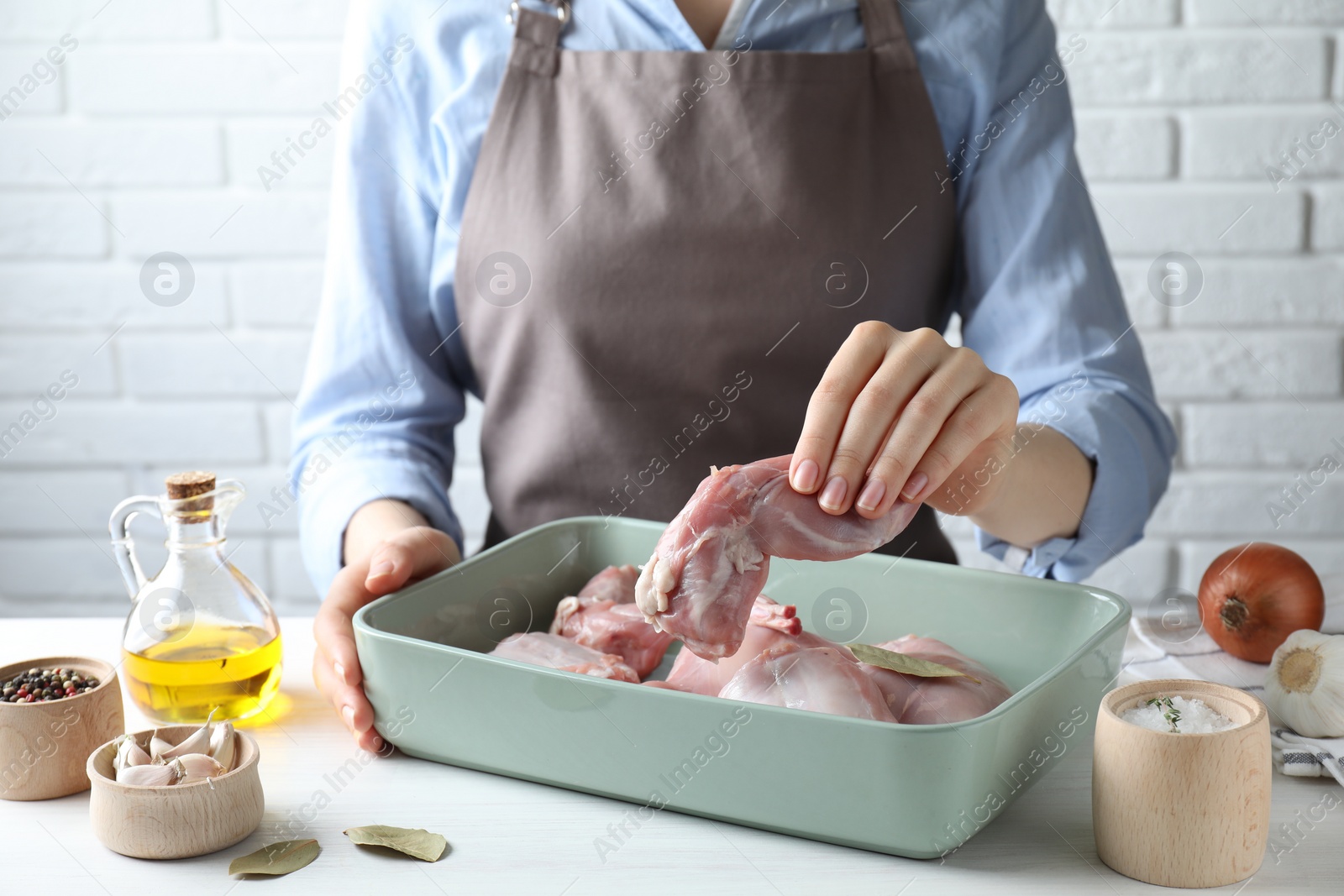 Photo of Woman putting rosemary into baking dish with raw rabbit meat at white wooden table, closeup
