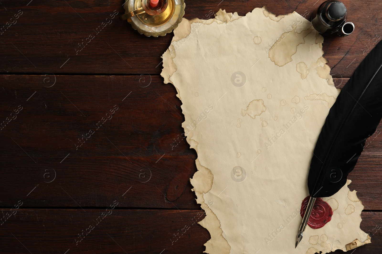 Photo of Sheet of old parchment paper with wax stamp, black feather, inkwell and candle on wooden table, flat lay. Space for text