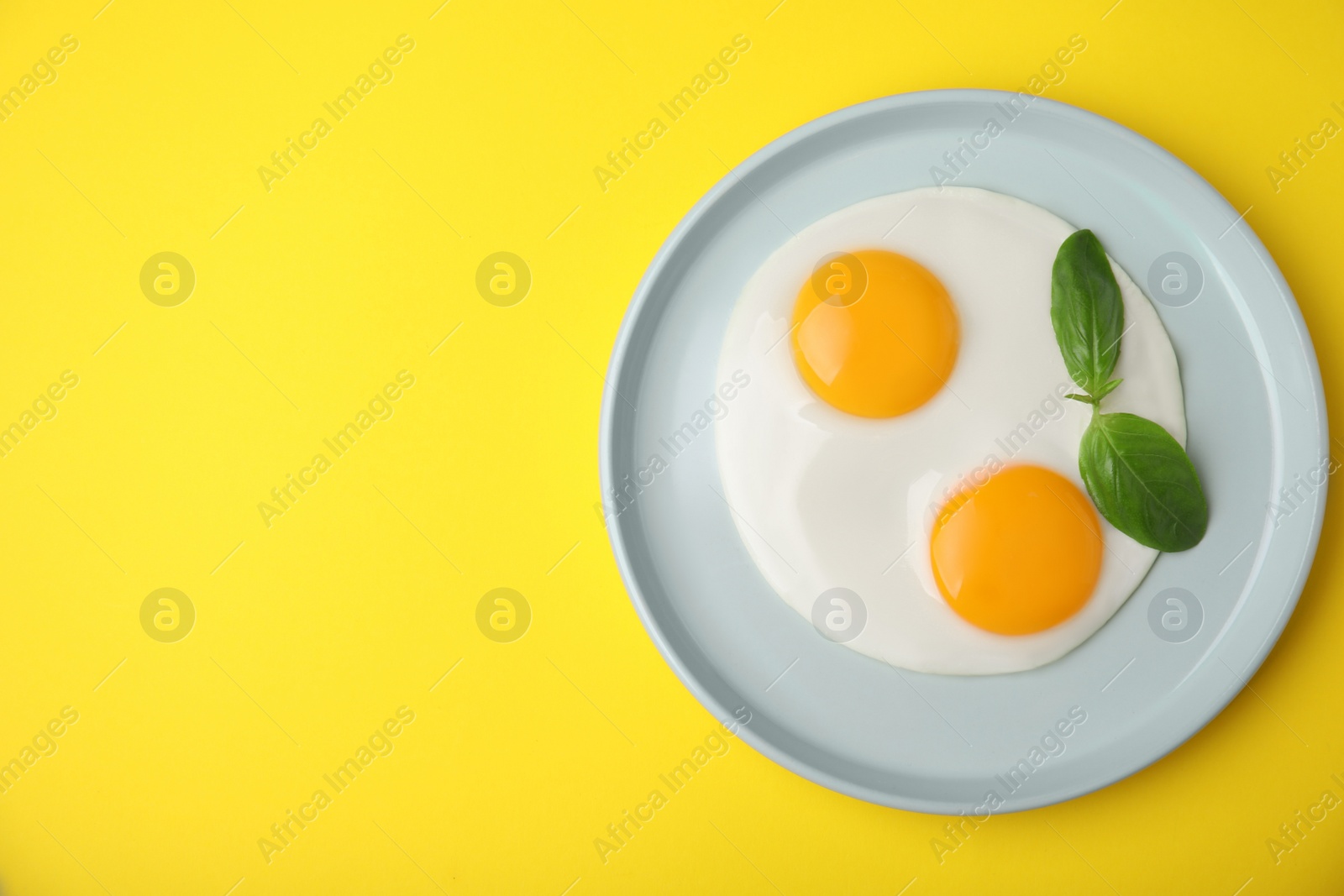 Photo of Tasty fried eggs with basil in plate on yellow background, top view. Space for text