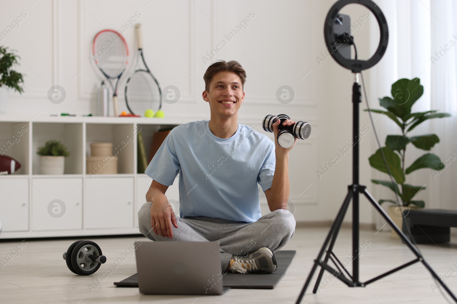 Photo of Smiling sports blogger holding dumbbells while streaming online fitness lesson at home