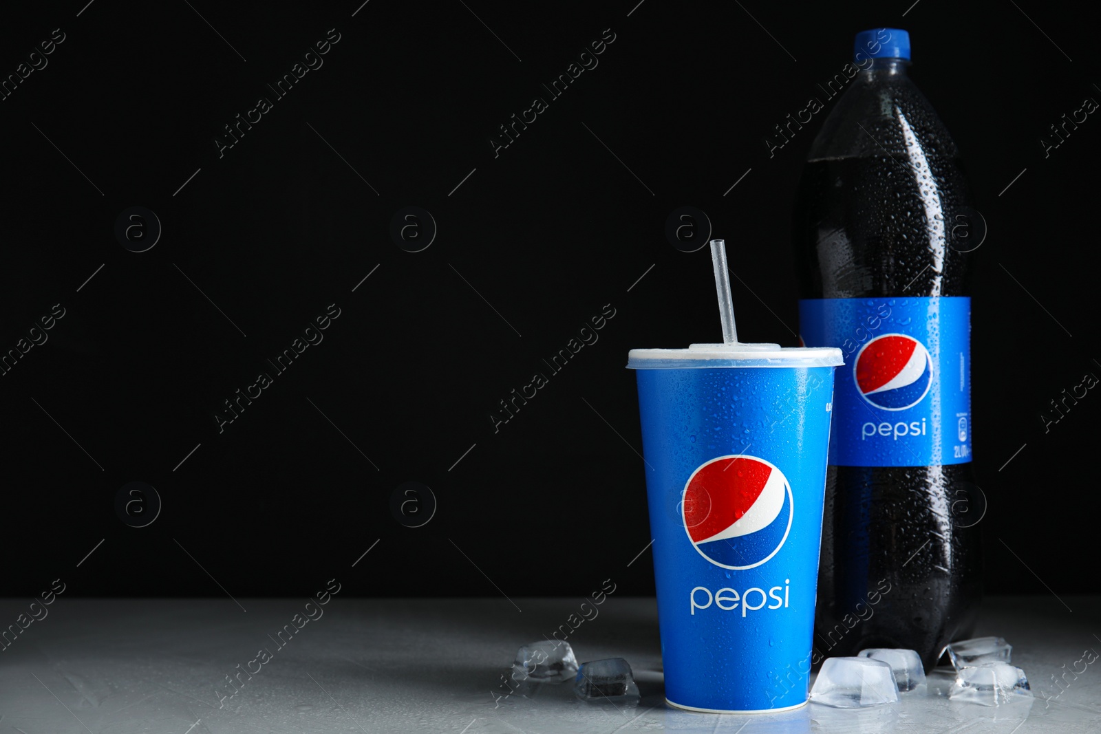 Photo of MYKOLAIV, UKRAINE - JUNE 9, 2021: Paper cup and bottle of Pepsi with ice cubes on light grey table against dark background, space for text