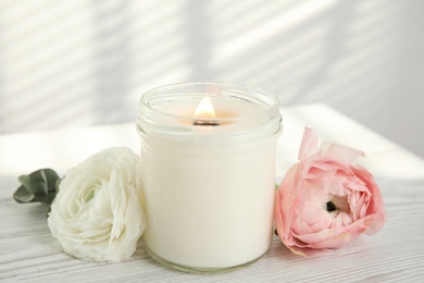 Candle with burning wooden wick and flowers on white table