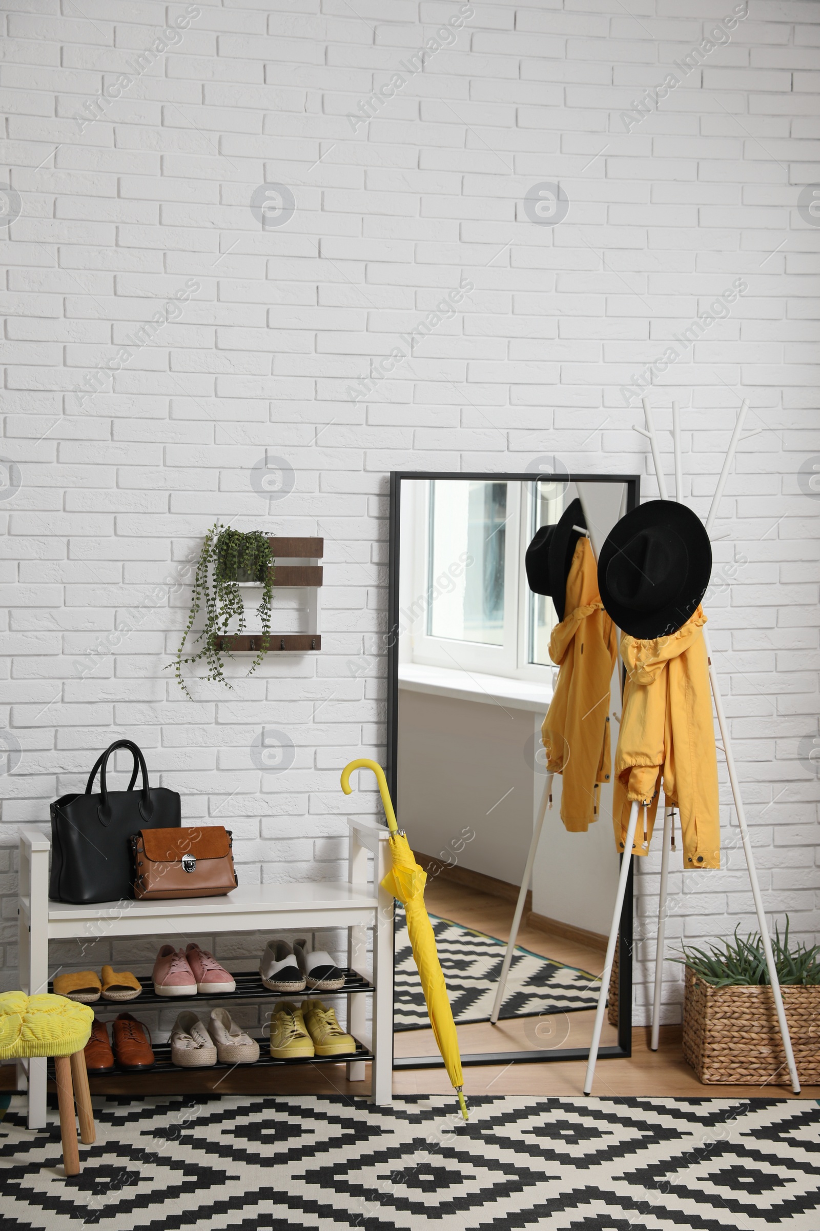 Photo of Stylish hallway interior with coat rack, shoe storage bench and mirror near white brick wall