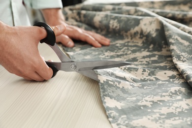 Photo of Professional tailor cutting camouflage fabric with scissors in workshop, closeup
